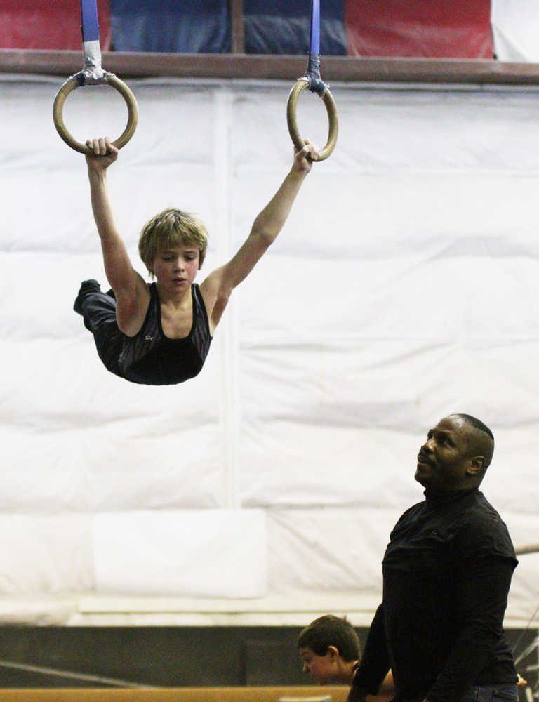 Caden Johns, of boys team level 5, works the still rings as coach Bill Dancil looks on.