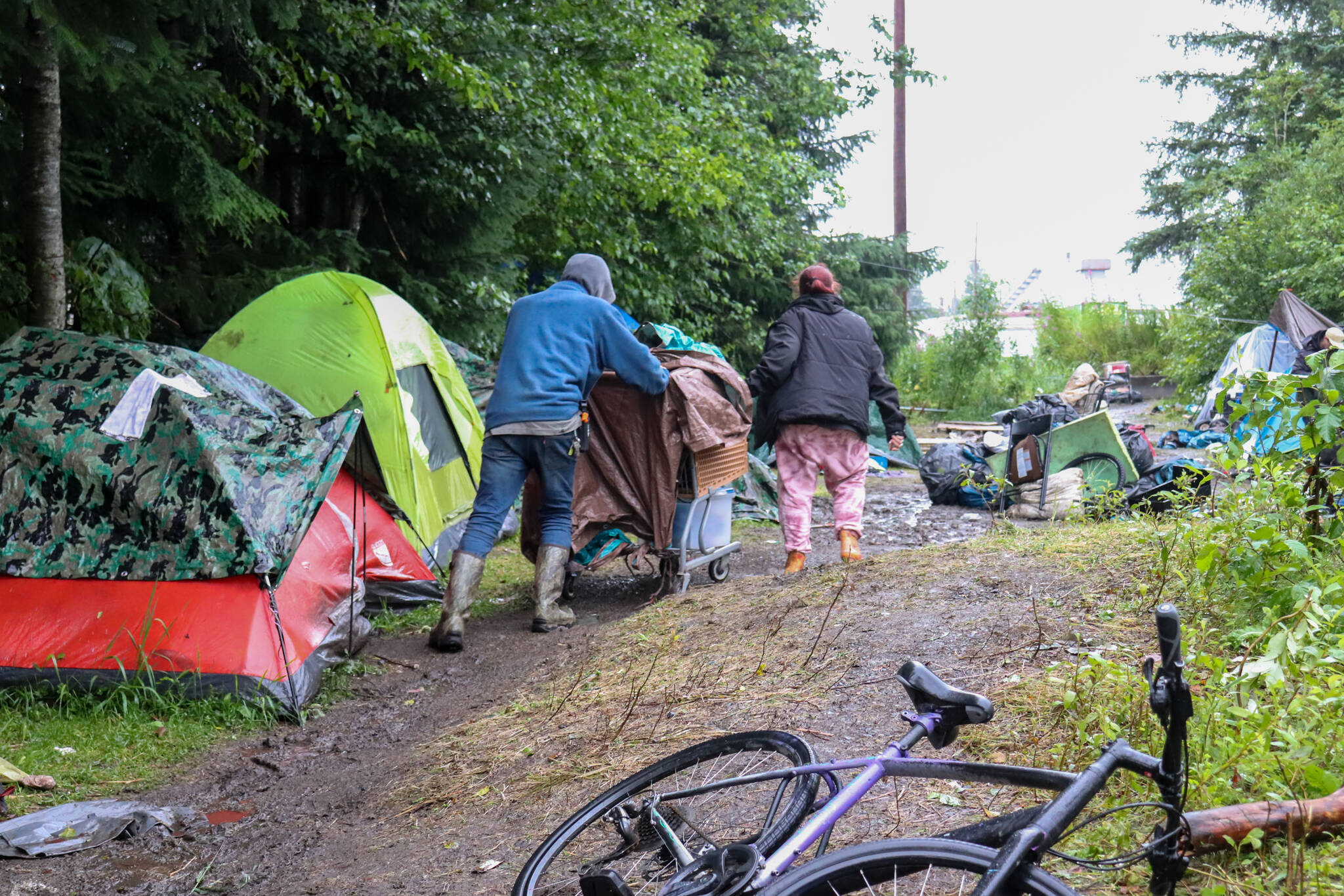 Christopher Moore helps another Juneau homeless resident wheel her belongings from a makeshift campsite on private property near the airport on Monday. (Jasz Garrett / Juneau Empire)
