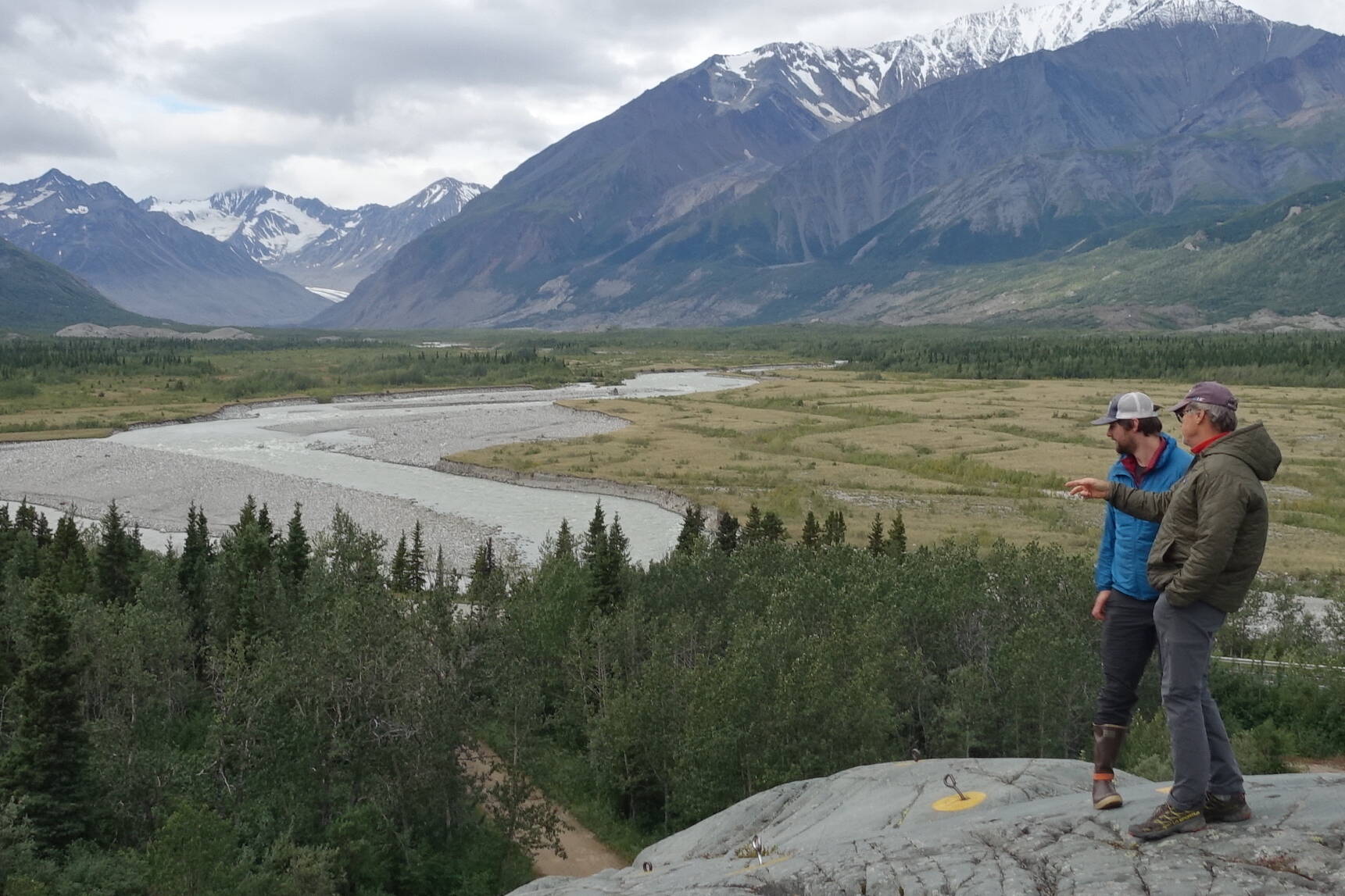 Phillip Wilson (blue jacket) and Dan Mann stand on a rock outcrop that was scoured by floodwaters a few centuries ago when Black Rapids Glacier — far in the distance — advanced to dam the Delta River. (Photo by Ned Rozell)