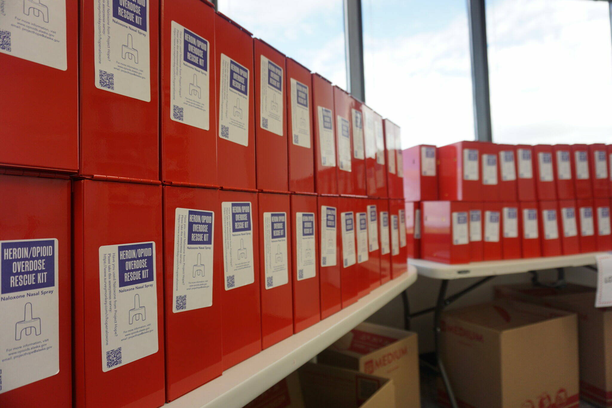 Boxed kits with naloxone, an overdose-reversal drug, and associated equipment are stacked on tables at the Alaska Department of Health’s Anchorage office on Aug. 9. The kits were assembled that day in preparation for distribution to school districts around the state, in accordance with House Bill 202. The bill requires schools to stock the kits and to have personnel trained to use them. (Yereth Rosen/Alaska Beacon)