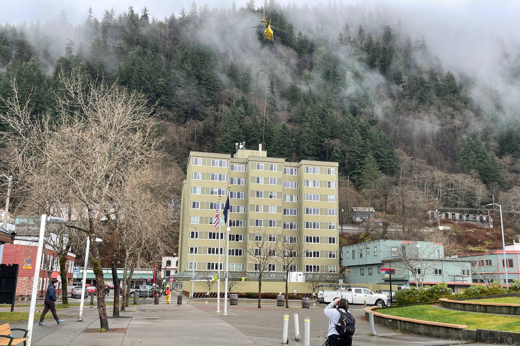 Aurelie Alexander photographs a helicopter hoisting cellular equipment onto the roof of the Marine View building at midday Wednesday. As a resident of the apartment/office building, she and others were notified to leave the building during the helicopter operation. (Laurie Craig / Juneau Empire)