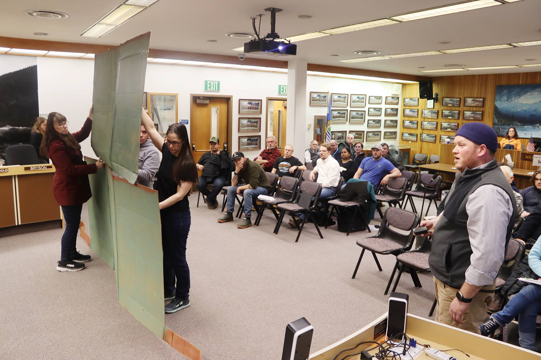 Noah Teshner (right) exhibits the physical impact military-grade flood barriers will have on properties with the help of other residents at a Juneau Assembly meeting on Monday night. (Mark Sabbatini / Juneau Empire)