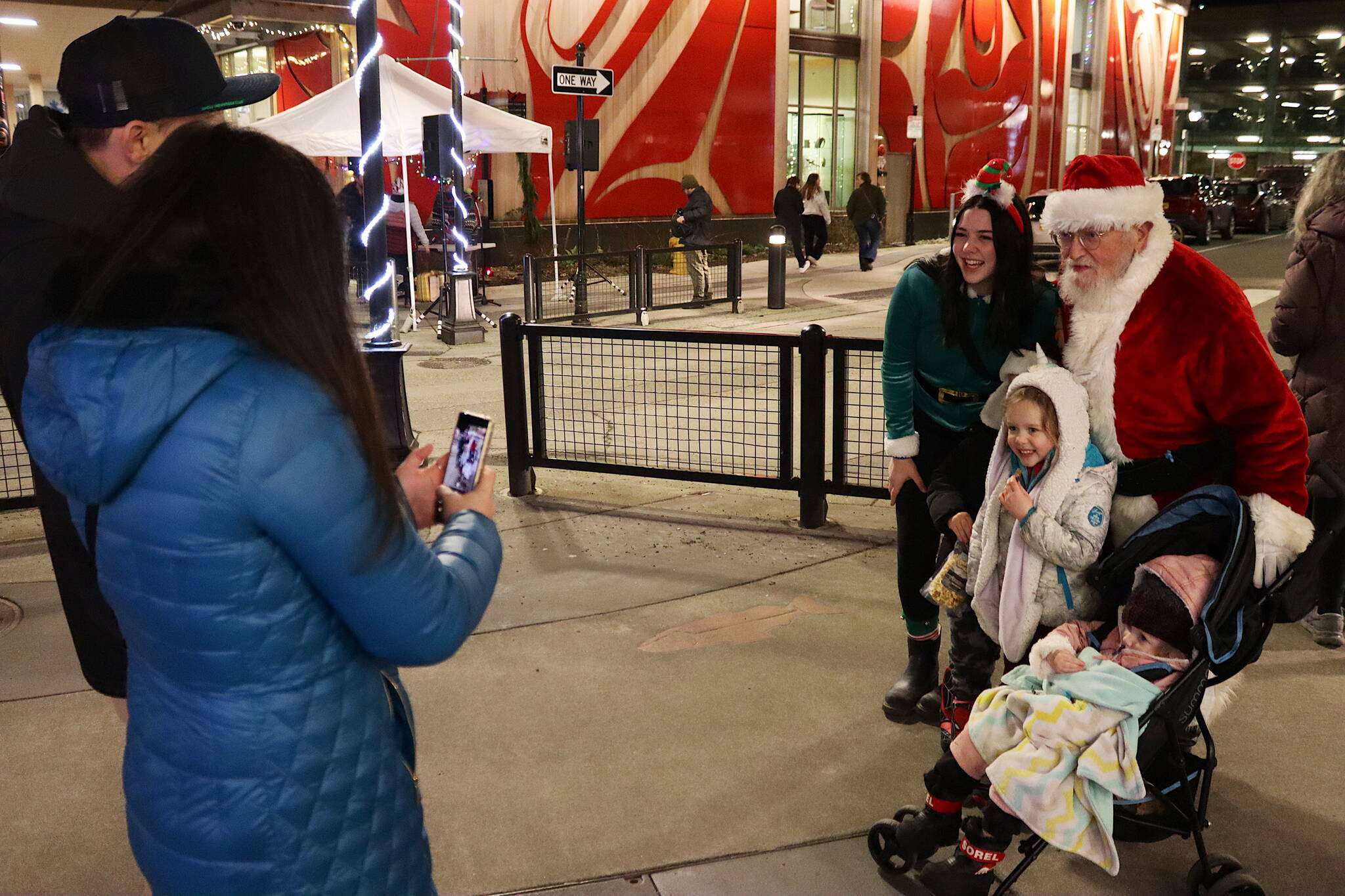 Marzena Whitmore (elf) and Dale Hudson (Santa), pose for a photo with Benny Orvin (partially obscured), 6, and his siblings Lilly, 4, and Remi, 2, taken by their mother Alex as their father Randy watches during last year’s Gallery Walk in downtown Juneau on Friday, Dec. 1, 2023. (Mark Sabbatini / Juneau Empire file photo)