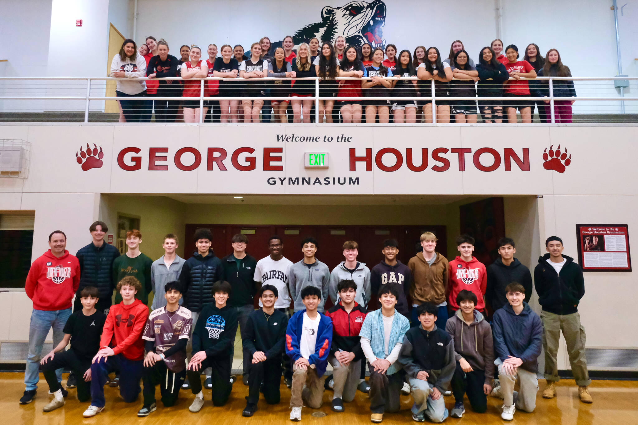 Members of the Juneau-Douglas High School: Yadaa.at Kalé Crimson Bears girls and boys basketball teams pose above and below the new signage and plaque for the George Houston Gymnasium on Monday. (Klas Stolpe / Juneau Empire)