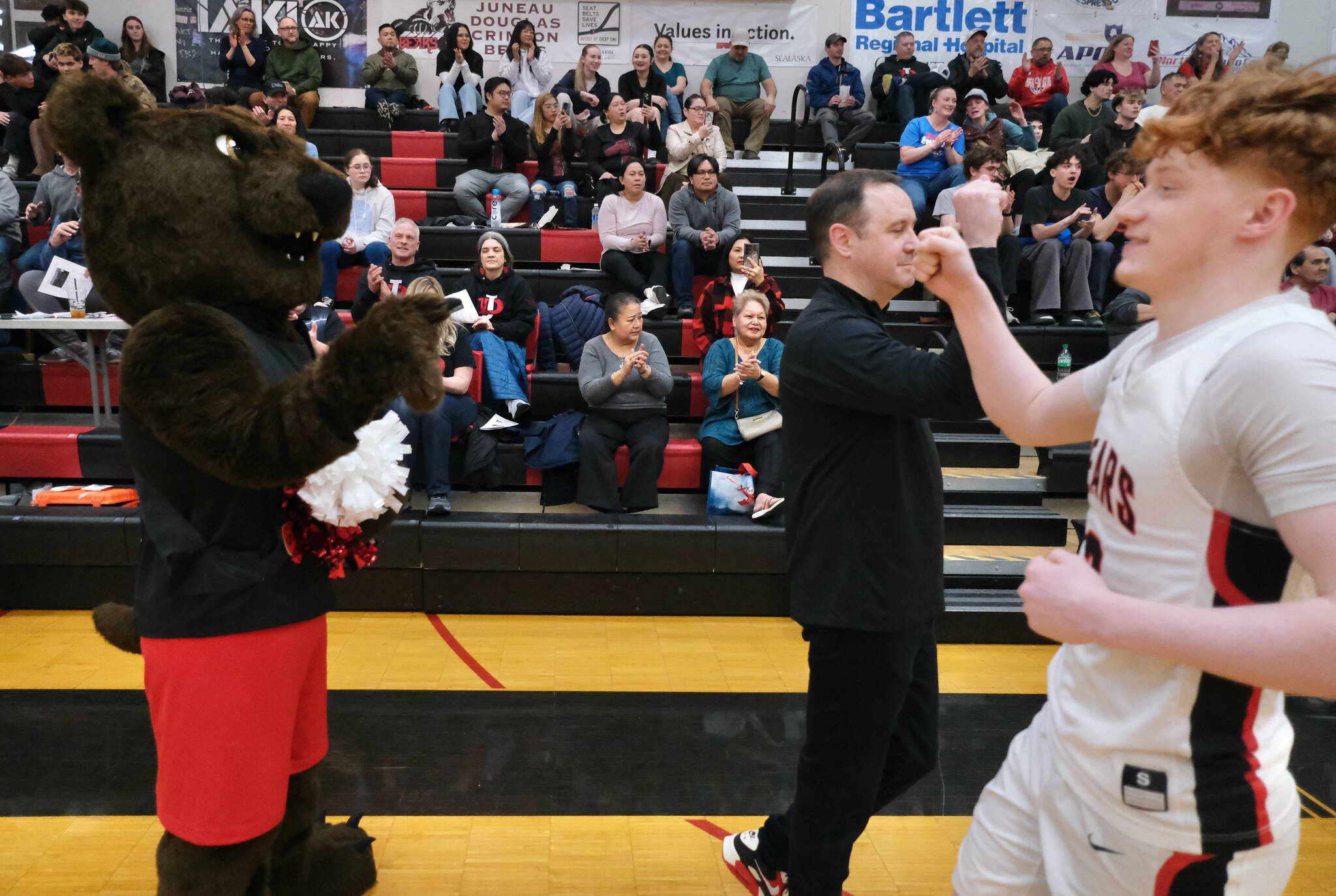 MAC the bear awaits a fist bump from Crimson Bears senior Gavin Gerrin after he runs past Juneau-Douglas High School: Yadaa.at Kalé coach Robert Casperson during Senior Night on Saturday at George Houston Gymnasium. (Klas Stolpe / Juneau Empire)